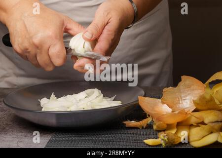 Gros plan des mains d'une femme dans un tablier blanc qui épluchent l'oignon avec un couteau Banque D'Images