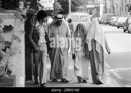 Des hommes arabes pauvres marchent dans la rue d'Earls court Londres 1970s Royaume-Uni. Les habitants du Moyen-Orient sont venus en Grande-Bretagne pour des soins de santé subventionnés dans les cliniques de Harley Street. Ils ont surtout séjourné dans le quartier d'Earls court. Quatre hommes, un jeune homme vêtu de l'Ouest et les trois autres hommes portant les robes blanches traditionnelles appelées un thoub, thobe, dishdasha ou kandora et la coiffure arabe traditionnelle appelée kaffiyeh ou ghutra. Earls court, Londres, Angleterre vers 1977 70s UK HOMER SYKES Banque D'Images