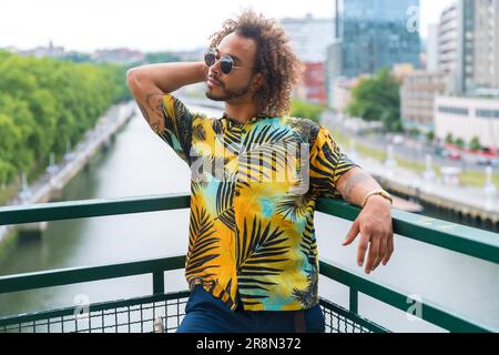 Passé d'un homme attrayant avec des cheveux afro en été portant une chemise de palmier et la ville en arrière-plan, en profitant de vacances Banque D'Images