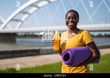 Femme sportive buvant une boisson énergétique et tenant un tapis d'exercice. Banque D'Images