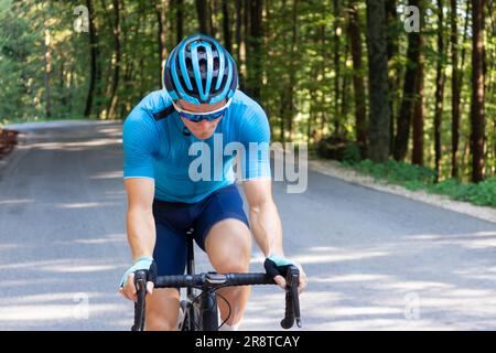 Cycliste de course automobile masculin dans un maillot de sport bleu, avec un casque et des lunettes, en montant une colline à travers la forêt. Banque D'Images