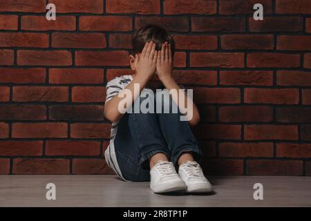 Garçon contrarié assis sur le sol près du mur de briques. Intimidation des enfants Banque D'Images