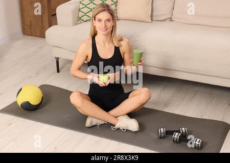 Jeune femme en vêtements de sport avec un verre de smoothie frais et de pomme à la maison Banque D'Images