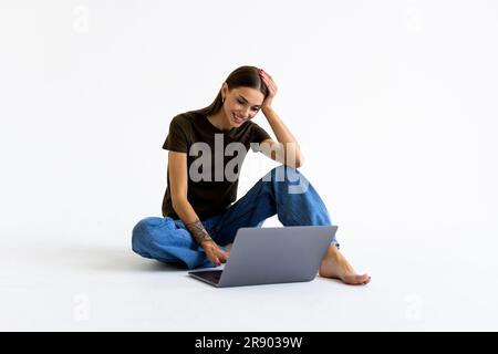 Portrait of a smiling casual girl holding laptop computer, assis sur un plancher isolé sur fond blanc Banque D'Images