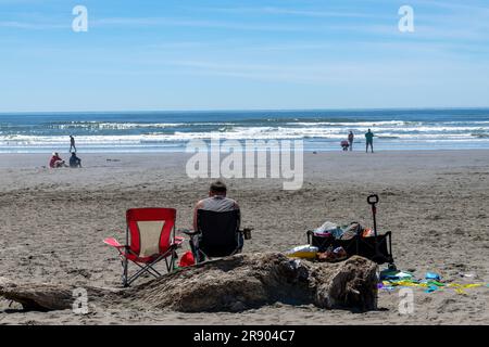 Vue sur la plage à l'océan d'un seul homme vu de l'arrière dans une chaise de plage contre un grand morceau de bois de drift se détendre avec quelques autres Beach-Goers dans le su Banque D'Images