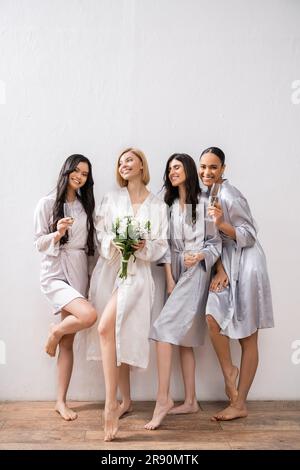 fête de mariée, amies interraciales tenant des verres avec champagne, mariée avec des fleurs blanches, femmes brunes et blondes, demoiselles, plongeur culturel Banque D'Images