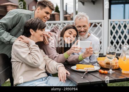 Une adolescente positive utilisant un smartphone près de parents matures et de père pendant la fête de barbecue avec nourriture d'été et fête de parents de jour à l'arrière-cour en juin Banque D'Images