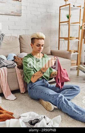 femme au style décontracté et tatouée avec une coiffure tendance assise sur le sol et triant des articles d'occasion près d'un canapé dans un salon moderne, durable Banque D'Images