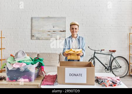 jeune et positive femme avec la coiffure tendance souriant à l'appareil photo et tenant le cavalier jaune près de la boîte de don et le contenant en plastique avec des vêtements, susta Banque D'Images