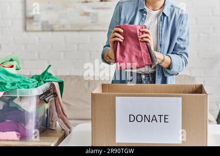 charité et bénévolat, vue rognée d'une jeune femme tatouée tenant des vêtements de garde-robe près de boîte en carton avec lettrage de don, vie durable Banque D'Images
