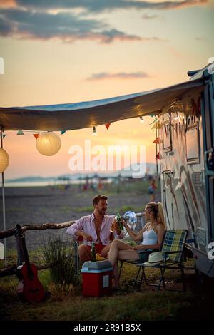 Jeune couple qui fête et toasque avec de la bière, assis devant le rv. Coucher de soleil en été. Voyage, vacances, ensemble, concept de style de vie. Banque D'Images