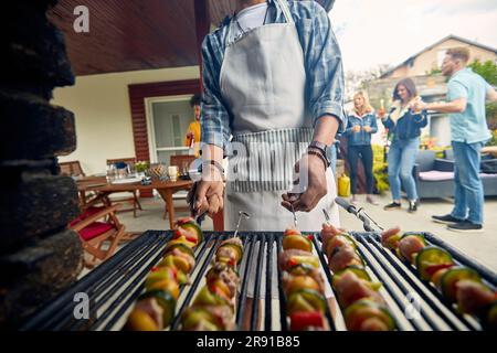 Vue rapprochée d'un gril qui grésilte. Au premier plan, les mains manœuvrent de manière experte des brochettes chargées de viandes et de légumes délicieusement marinés sur le Banque D'Images