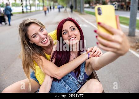 Une femme heureuse avec un ami qui prend le selfie par téléphone portable sur banc Banque D'Images