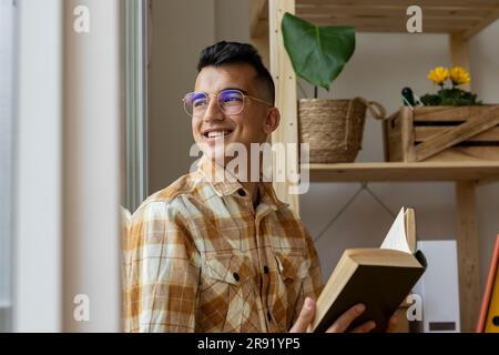 Un homme heureux avec un livre qui regarde par la fenêtre à la maison Banque D'Images