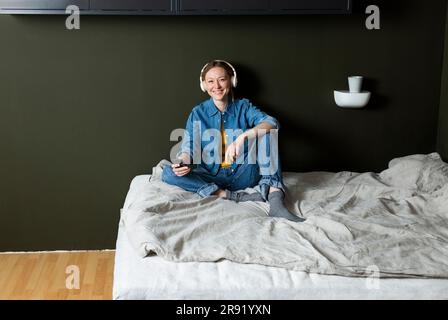 Jeune femme souriante qui écoute de la musique avec un casque sur le lit Banque D'Images