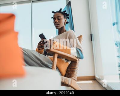 Jeune femme utilisant un smartphone assis dans un fauteuil à la maison Banque D'Images