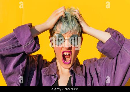 Femme avec les mains dans les cheveux criant devant le mur jaune Banque D'Images