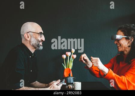 Homme souriant avec une femme portant un casque de réalité virtuelle devant un mur noir Banque D'Images