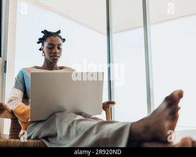 Jeune femme utilisant un ordinateur portable assis confortablement dans un fauteuil à la maison Banque D'Images