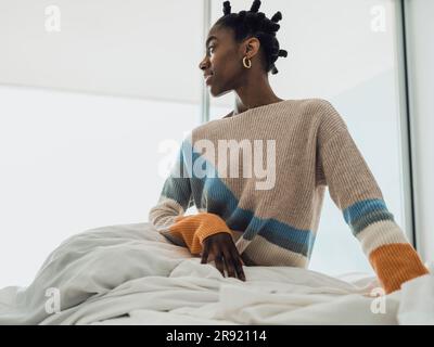 Une jeune femme heureuse assise sur le lit et qui regarde loin Banque D'Images