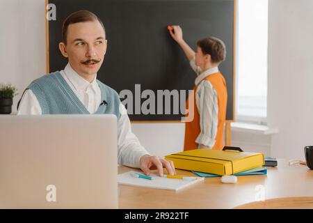 Homme souriant assis dans une salle de classe regardant un ordinateur portable tandis que son collègue écrit sur un tableau noir Banque D'Images