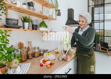 Femme souriante gardant le sac en filet plein de provisions sur le comptoir dans la cuisine à la maison Banque D'Images