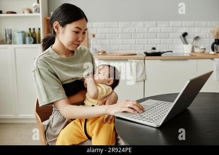 Une femme indépendante souriante qui allaite sa fille et utilise son ordinateur portable à la maison Banque D'Images