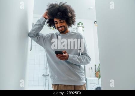 Homme souriant avec la main dans les cheveux à l'aide d'un smartphone dans la salle de bains à la maison Banque D'Images