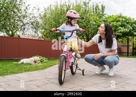 Une mère souriante se caroupe par sa fille en apprenant à monter à vélo sur une piste de marche Banque D'Images
