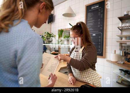 Propriétaire de café avec le syndrome de Down aidant le client à choisir la nourriture dans le menu dans le café-restaurant Banque D'Images