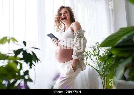 Femme enceinte heureuse avec main dans la poche tenant un smartphone à la maison Banque D'Images