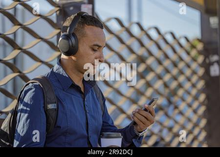 Plan moyen d'un jeune Latino-américain avec un casque regardant son téléphone portable à l'arrêt de bus vu de derrière. Banque D'Images