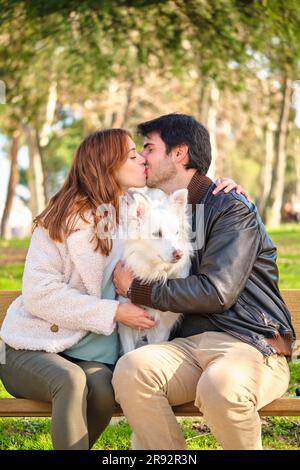 Un jeune couple caucasien embrasse son chien assis sur un banc dans un parc. Famille. Banque D'Images