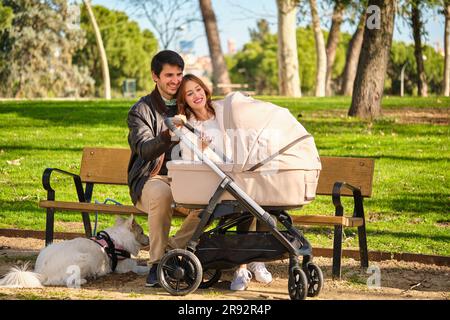 Couple regardant leur bébé dans une poussette de bébé avec leur chien assis sur un banc dans un parc. Banque D'Images