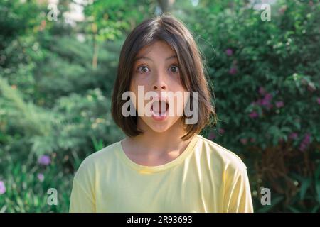 Portrait en gros plan d'une adolescente très surprise avec de larges yeux ouverts et la bouche sur le fond de verdure. Banque D'Images