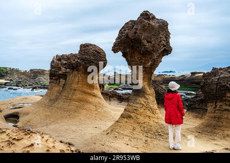 Yehliu Geopark, Taïwan -- 14 mars 2023. Une femme touriste dans un manteau rouge vif et chapeau blanc se tient près d'une formation de roche typiquement étrange. Banque D'Images