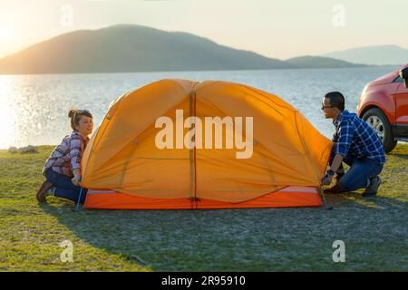 Couple asiatique préparant une tente pour camper dans la pelouse avec le lac en arrière-plan pendant le coucher du soleil Banque D'Images