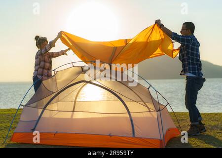 Couple asiatique préparant une tente pour camper dans la pelouse avec le lac en arrière-plan pendant le coucher du soleil Banque D'Images