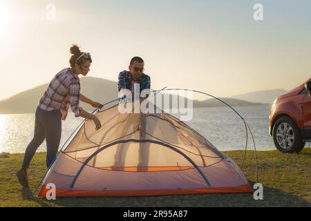 Couple asiatique préparant une tente pour camper dans la pelouse avec le lac en arrière-plan pendant le coucher du soleil Banque D'Images