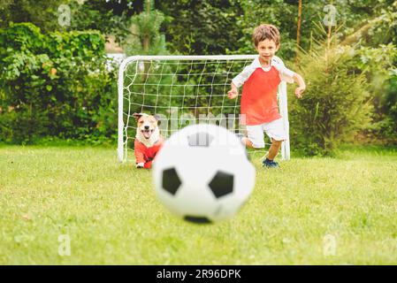 Joyeux enfant dans un kit de football jouant au football avec un chien de compagnie de famille sur la pelouse du jardin le jour d'été Banque D'Images