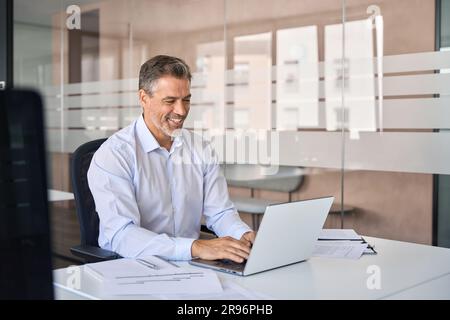 Homme d'affaires souriant d'un âge moyen, cadre assis au bureau à l'aide d'un ordinateur portable. Banque D'Images