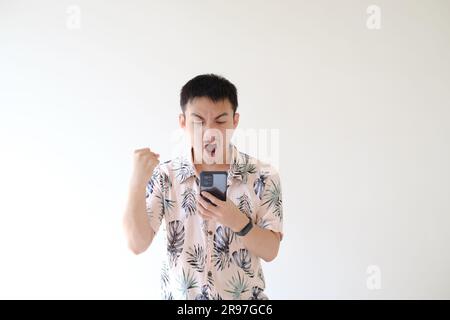 Un jeune asiatique portant une chemise rose aux motifs tropicaux et une montre intelligente sur son poignet gauche est excité en regardant son smartphone. Isolé Banque D'Images