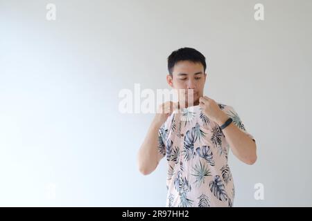 Un jeune homme asiatique portant une chemise rose aux motifs tropicaux et une montre intelligente sur son poignet gauche souffle l'intérieur de la chemise à cause de la hea Banque D'Images