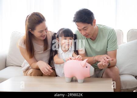 Père de famille asiatique heureux et mère et fille assis sur canapé planification de financement avec des économies tout en déposant des pièces dans la chambre, maman et papa Banque D'Images