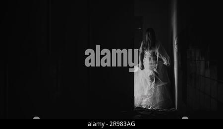 Scène d'horreur d'une mariée zombie possédée, femme en robe blanche fantôme tenant des roses séchées dans la pièce sombre. Halloween effrayant concept Banque D'Images