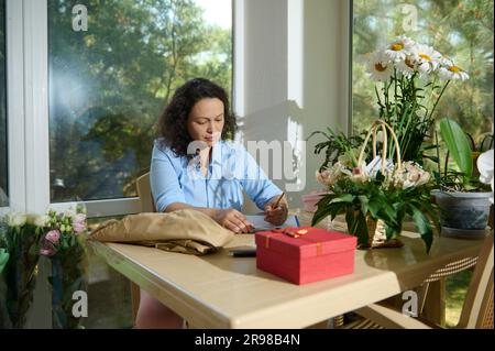 Fleuriste féminine dessinant sur le presse-papiers une composition de fleurs pour un événement de vie spécial, assis à table dans un fleuriste Banque D'Images