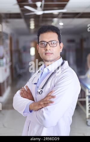 Portrait de médecin masculin souriant en blouse de laboratoire avec les bras croisés, médecin indien heureux en blouse blanche souriant dans le couloir de l'hôpital. Banque D'Images