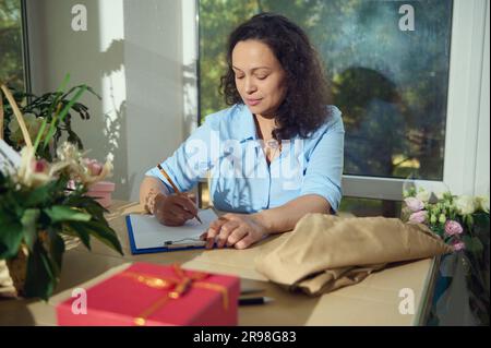 Femme afro-américaine enceinte, propriétaire d'entreprise travaillant à la boutique florale, recevant la commande, écrivant des notes sur le presse-papiers Banque D'Images