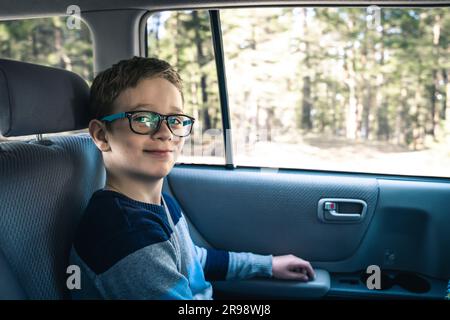 Un garçon caucasien heureux d'âge scolaire se déplace dans le siège arrière d'une voiture et regarde la caméra . Un écolier avec des verres. L'enfant porte la ceinture de sécurité Banque D'Images