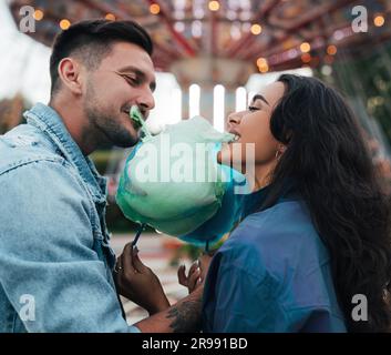 Jeune couple heureux piquant des bonbons en coton. L'homme et sa petite amie mordent un bonbon de coton bleu contre le carrousel. Banque D'Images